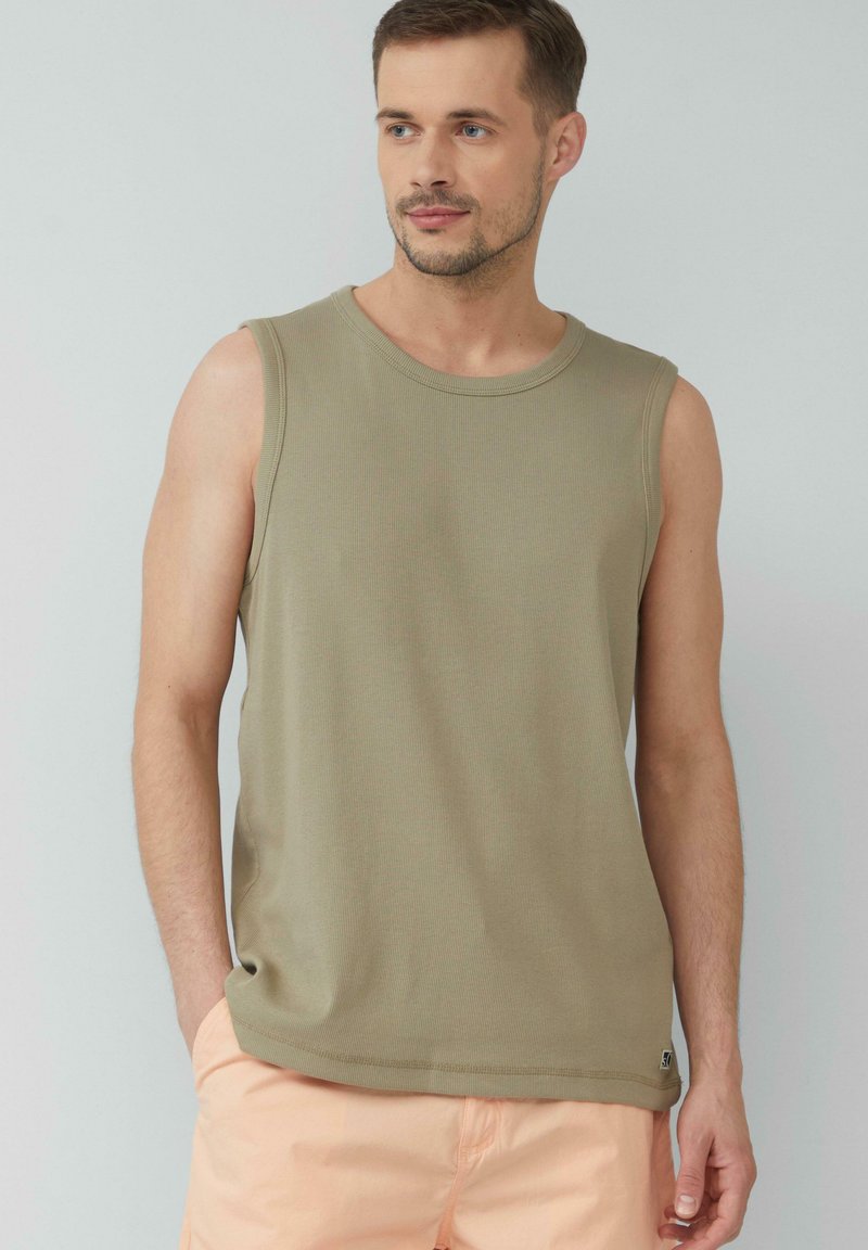 Olivegrünes, ärmelloses Tanktop aus gerippter Textur, mit Rundhalsausschnitt und dezentem Logo-Detail am Saum. Getragen mit hellpfirsichfarbenen Shorts.
