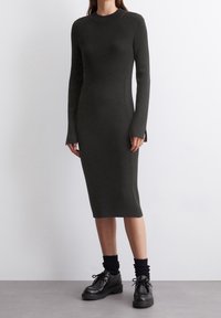 Robe côtelée à manches longues, de couleur vert foncé, avec une silhouette ajustée et des fentes sur les côtés. Associée à des chaussures noires à lacets et des chaussettes noires texturées.