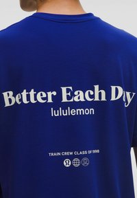 Kék póló fehér szöveggel: "Jobb Minden Nap" és "lululemon", valamint "Edzői Osztály 1998" és három kis logó a szöveg alatt.