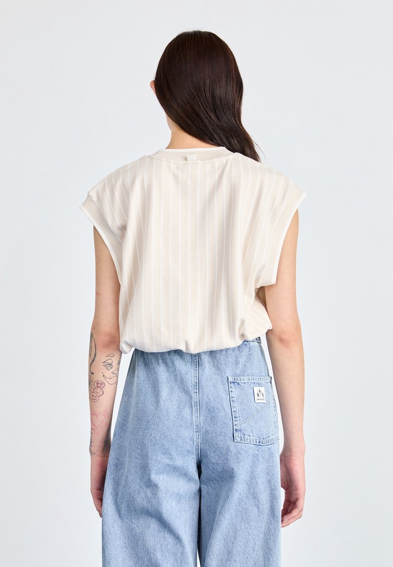 Person med långt mörkt hår bär en ärmlös beige randig topp och högmidjade ljusblå denimjeans, sedd bakifrån.