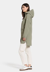 Oljgrön vattentät jacka med huva, tryckknappar, elastisk midja och sidospalter, kombinerad med krämfärgade byxor och vita sneakers.