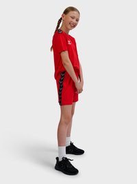 Smilende pige med fletninger iført et rødt sportstøj med sorte sidelister, hvide sokker og sorte sneakers, står sidelæns.