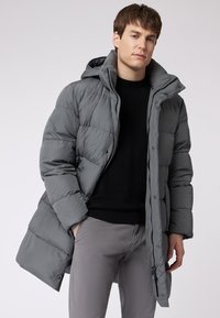 Grå pufferjakke med hætte, med en quiltet tekstur, trykknapper og sidelommer. Båret over en sort sweater og lysegrå bukser.