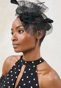 Crni fascinator od tila s mrežastim uzorkom, ukrašen mašnom i perjem. Nošen uz haljinu s točkicama i naušnicama na obruče.