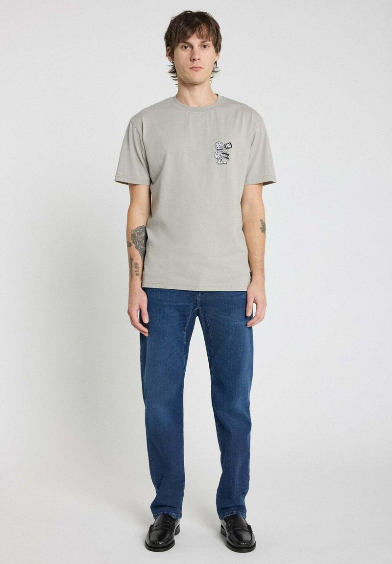 T-shirt en coton gris avec un motif imprimé sur la poitrine gauche, associée à un jean bleu. Le modèle se tient devant un fond uni.