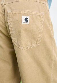 Béžové kordové nohavice s príklopkovou vreckom, na ktorom je biely štítok s logom Carhartt. Textúra je rebrovaná, čím zvýrazňuje odolnosť.