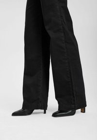 Jean noir à jambes larges avec un ourlet brut, associé à des talons pointus noirs à petit talon carré. Les matériaux semblent lisses et résistants.