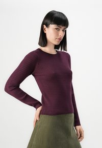 Pull en maille côtelée bordeaux à manches longues, avec un décolleté rond et une coupe ajustée, associé à une jupe verte texturée.