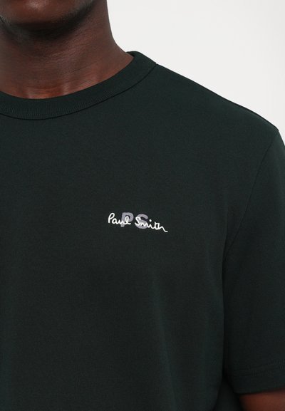 Vyras dėvi tamsiai žalią marškinėlį su mažu baltu ir pilku "Paul Smith PS" logotipu kairėje krūtinės pusėje, kirptu ties kaklu ir viduriniu liemeniu.