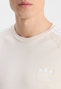 Beige t-shirt med kort ärm, tillverkad av ett mjukt tyg, med rund halsringning och vit tre-stremsdesign på axlarna med broderad logotyp.