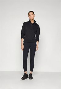 Donna in piedi contro un muro grigio, che indossa un pullover nero con zip, leggings neri e scarpe da ginnastica nere, rivolta in avanti con una postura rilassata.