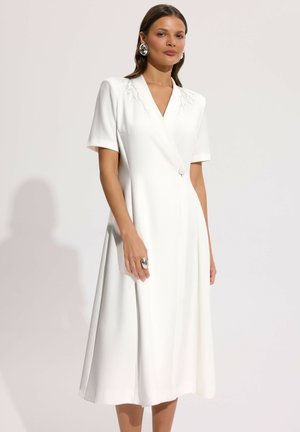 Kobieta w białej sukience midi z krótkimi rękawami, zdobieniami na ramionach i pojedynczym guzikiem, ozdobiona dużymi srebrnymi kolczykami i pierścionkiem.