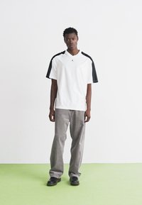 Camiseta deportiva blanca con acentos negros en los hombros, combinada con pantalones anchos grises y zapatos negros, sobre un fondo claro.