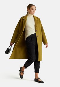 Olijfgroen wollen jas met ingesneden revers, bruine knopen, gecombineerd met een roomkleurige coltrui en zwarte corduroy broek. Gecombineerd met een kleine zwarte handtas.