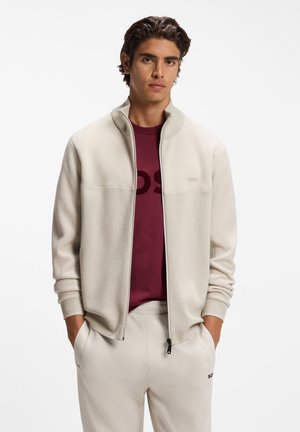 Junger Mann trägt eine cremeweiße Jacke mit Reißverschluss über einem dunkelroten Hemd und passenden cremeweißen Hosen, die Hände in den Taschen, stehend vor weißem Hintergrund.