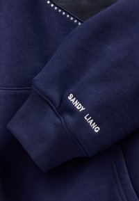 Granatowa bluza z teksturowanej tkaniny, z subtelnym błyszczącym akcentem z kryształków na ramieniu oraz haftowanym napisem „SANDY LIANG” na mankiecie.