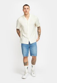 Linnen korte mouwen shirt in gebroken wit, gecombineerd met blauwe denimshorts. Heeft een knoopsluiting, casual pasvorm en zichtbare tatoeages op de armen.