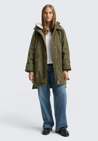 Parka verde oliva con cappuccio foderato in pelliccia, chiusura con zip e ampie tasche laterali, abbinato a jeans a gamba larga azzurri e scarpe con plateau nere.