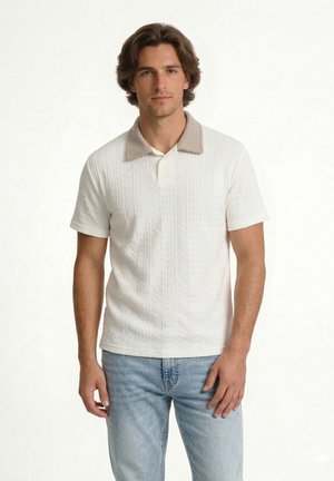 Mand med mellem langt brunt hår iført en hvid kortærmet tekstureret polo med beige krave og lyseblå jeans, stående mod en hvid baggrund.