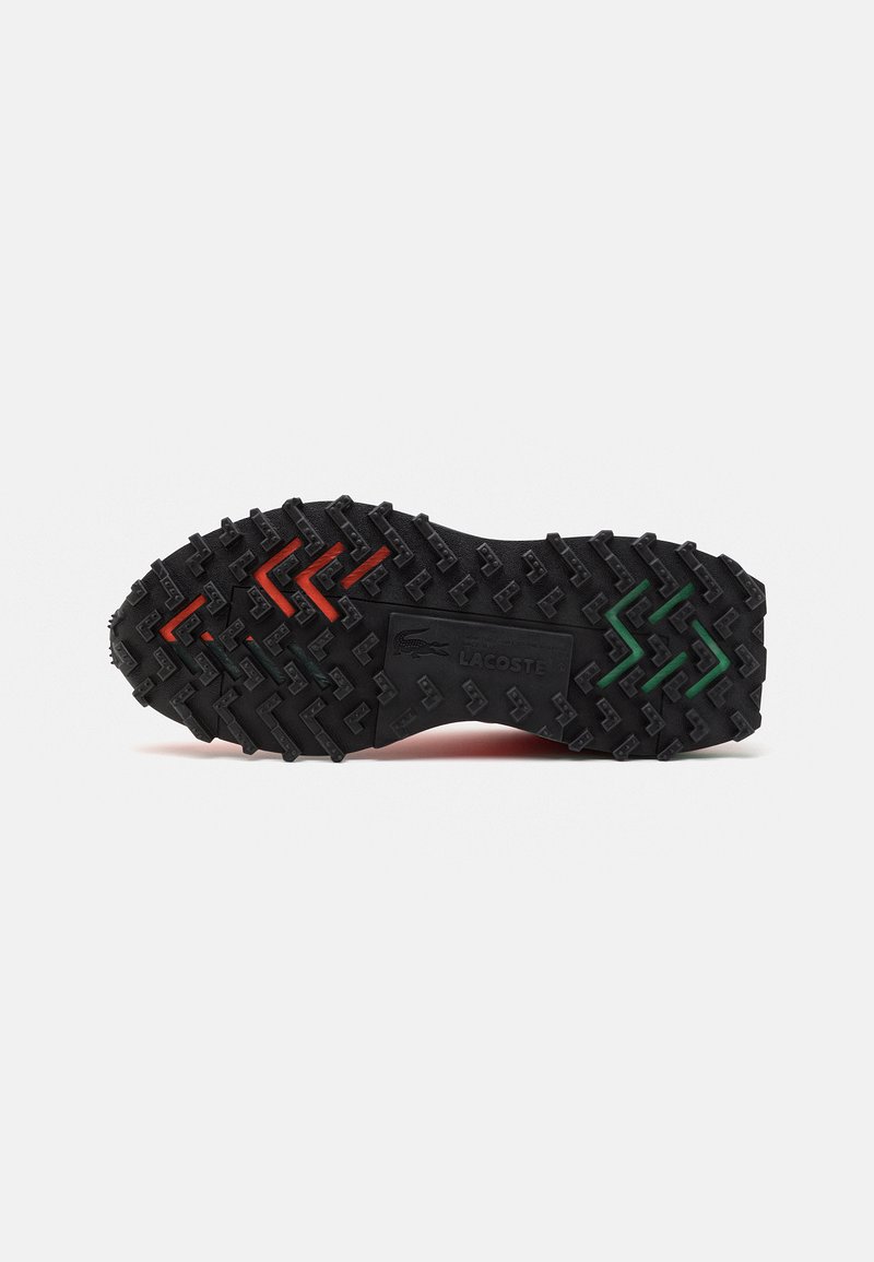 Suela de goma negra con textura; presenta acentos en zigzag en rojo, verde y naranja. Marca Lacoste visible en el lateral.
