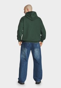 Zielona bluza z kapturem, posiadająca kieszeń kangurkę z przodu oraz sznurek ściągający, w połączeniu z luźnymi, wyblakłymi niebieskimi dżinsami z tylnymi kieszeniami.