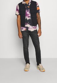 Czarna kamizelka z wieloma kieszeniami na fioletowo-białej koszulce z krótkim rękawem z efektem tie-dye, połączona z ciemnymi dżinsami i jasnymi tenisówkami.