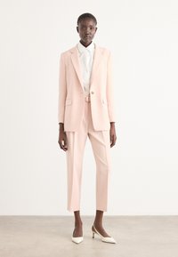 Completo su misura rosa chiaro con giacca strutturata; pantaloni attillati; tessuto testurizzato; abbinato a una camicia bianca e scarpe con tacco bianche.