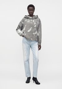 Άτομο που φοράει γκρι φούτερ tie-dye, ανοιχτά μπλε τζιν με πιτσιλιές μπογιάς και μαύρα μποτάκια μέχρι τον αστράγαλο, στέκεται σε απλό φόντο.