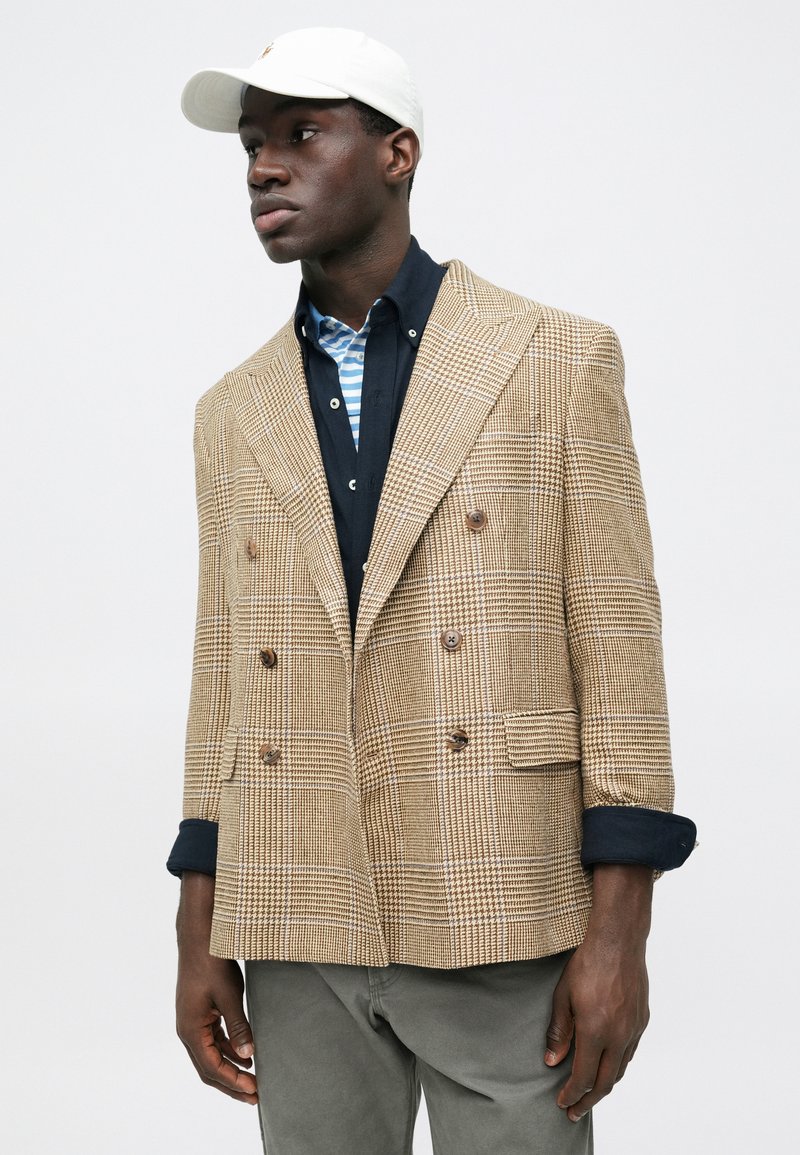 Brown plaid double-breasted blazer with large lapels, textured fabric, and brown buttons. Worn over a navy shirt and grey pants.