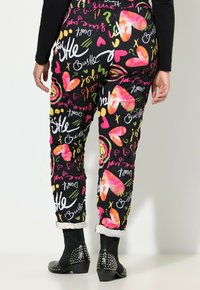 Persoon in zwarte broek met kleurrijke harten, tekstzinnen en opgerolde broekspijpen, gecombineerd met zwarte studs enkelboots, gezien van achteren.