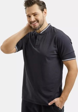 Uśmiechnięty mężczyzna z brodą w ciemnogranatowej koszulce polo z białymi paskami na kołnierzyku i krawędziach rękawów, pozuje z ręką za szyją.