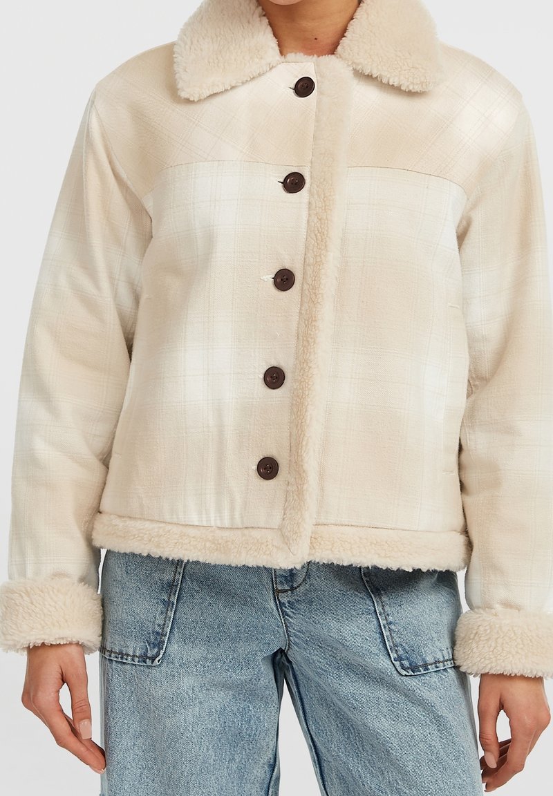 Veste beige doublée en sherpa avec boutons foncés, portée avec un jean denim bleu clair, les mains détendues le long du corps.