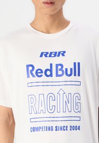 T-shirt en coton blanc avec un texte imprimé bleu : "RBR Red Bull RACING" et "COMPÉTITION DEPUIS 2004", avec un design simpliste et un col rond.