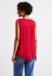 Blusa rossa senza maniche in tessuto morbido, con dettaglio arricciato sul rétro, orlo curvo e vestibilità rilassata, abbinata a jeans blu chiaro.