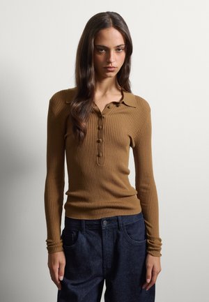Junge Frau mit langen braunen Haaren, die ein braunes, geripptes Langarmshirt und dunkelblaue Jeans trägt, stehend vor einem einfachen weißen Hintergrund.