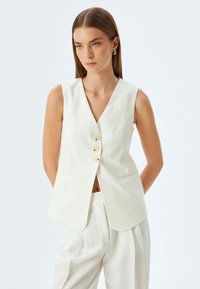 Witte linnen mouwloze vest met een V-hals, drie knopen aan de voorkant en twee zijzakken. Gecombineerd met lichtgestreepte broek.