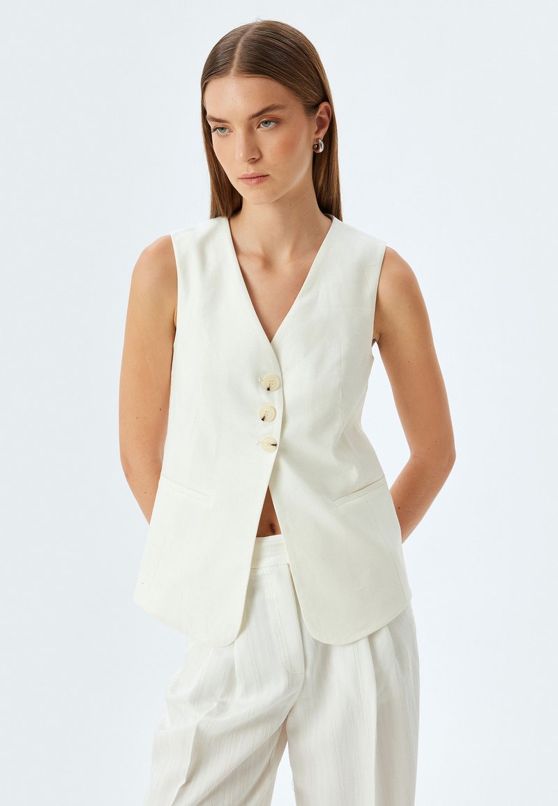 Witte linnen mouwloze vest met een V-hals, drie knopen aan de voorkant en twee zijzakken. Gecombineerd met lichtgestreepte broek.