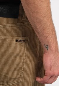 Brune fløjl bukser med en tekstureret overflade, sidelomme med et sort og orange "VOLCOM" label og en synlig tatovering på håndleddet.
