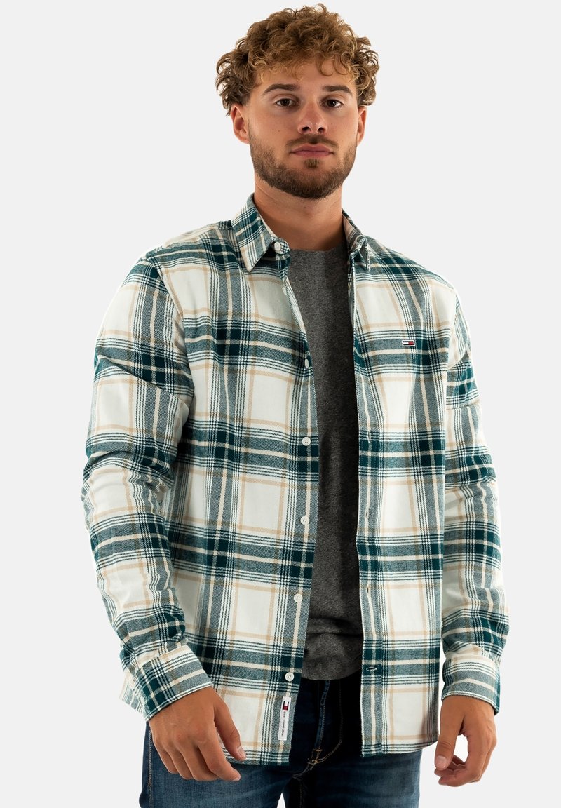 Camisa de franela a cuadros en azul, verde y beige sobre una base blanca, de manga larga, con bolsillos delanteros y cierre de botones. Usada sobre una camiseta gris.