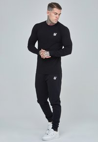 Black long-sleeve shirt and matching joggers made of a smooth fabric, featuring a small white logo on the chest and pants. White sneakers.