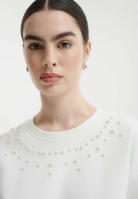 Bílá mikina s texturovaným designem a malými ozdobnými perlovými akcenty uspořádanými podél výstřihu a ramen.