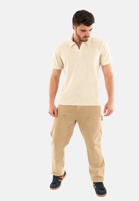 Polo beige con colletto, abbinato a pantaloni cargo beige con più tasche. Calzature: sneakers blu navy con strisce arancioni.