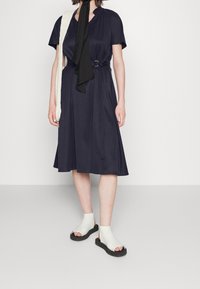 Mujer con un vestido negro marino con cinturón a la altura de la rodilla, una bufanda negra en el cuello, sandalias blancas de punta abierta con suelas negras y un bolso de hombro blanco texturizado.