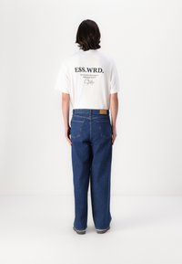 Tricou alb cu text pe spate, asociat cu blugi largi de culoare albastră. Denim-ul are două buzunare îndărăt și o etichetă din piele bej pe talie.