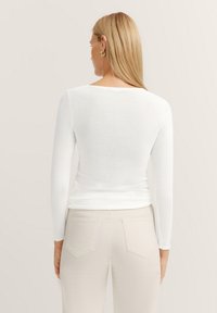 Weißes Langarmoberteil mit rundem Ausschnitt, glatter Textur und figurbetontem Design, kombiniert mit hellbeigen Hosen mit Gesäßtaschen.