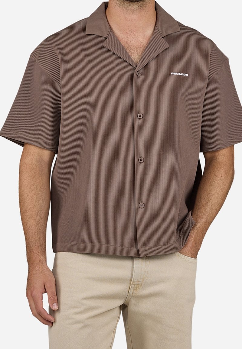 Homme portant une chemise marron côtelée à manches courtes, boutonnée avec un col, associée à un pantalon beige clair, main dans la poche.