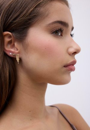 Jeune femme de profil portant trois boucles d'oreilles en or : un clou de perle, un cœur et un modèle à franges en chaîne, avec de longs cheveux bruns.