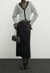 Cardigan en tricot rayé blanc et marine, jupe en satin noire, sac à main vert olive et sandales noires à lanières, se tenant sur une surface texturée.