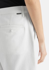Pantaloni di cotone grigio chiaro con una texture liscia, dotati di una tasca posteriore con chiusura a pulsante e di un piccolo etichetta del marchio sul cinturino.