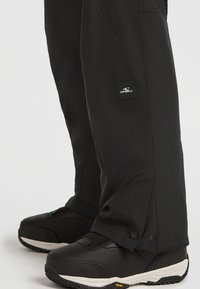 Zwarte sneeuwbroeken met een gestructureerd oppervlak en een klein O'Neill logo op het label, gecombineerd met stevige zwarte boots met een witte zool en accenten.
